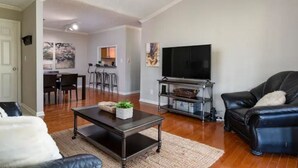 A living room with a black leather sofa, a dark wooden coffee table, and a flat-screen TV on a stand. There is a dining area with a table and chairs in the background.