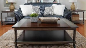 A living room with a dark leather sofa, two white pillows, a wooden coffee table, a small potted plant, and a white shelving unit with decorative items.