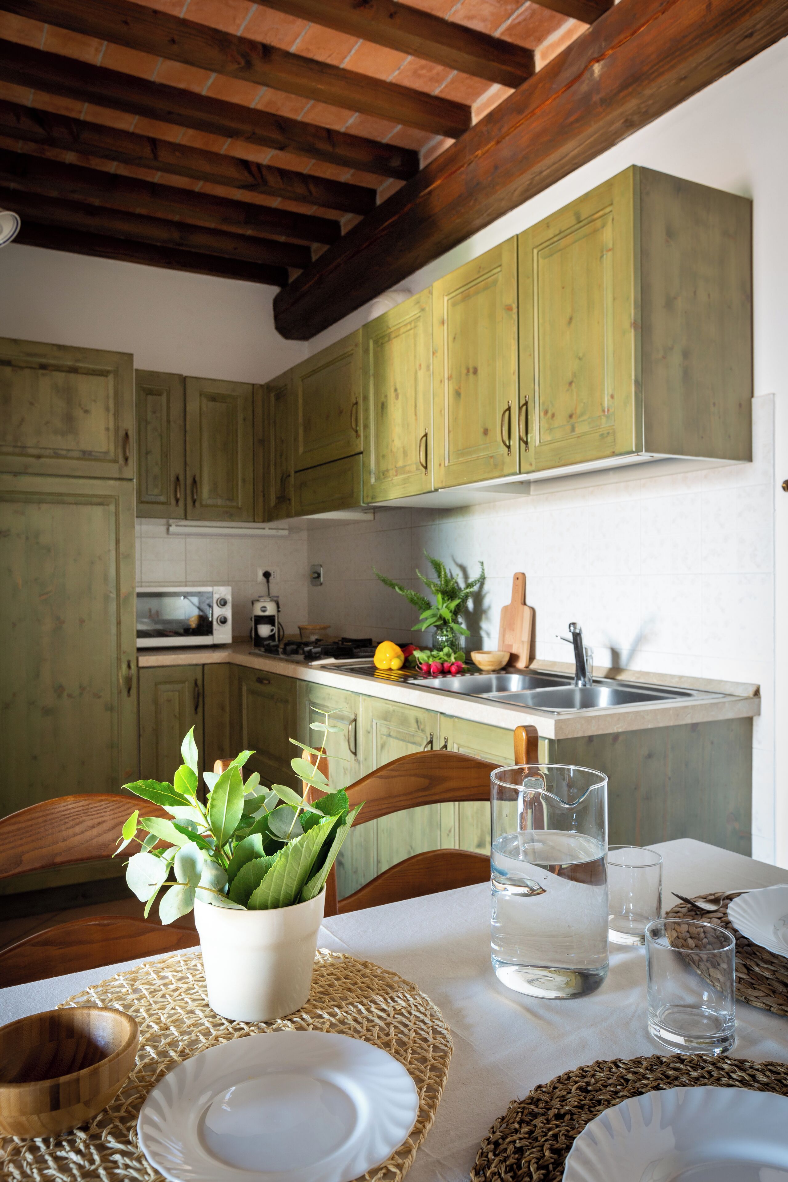 A rustic kitchen with wooden beams, green cabinets, and a dining area with a table set for two.