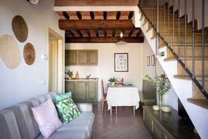 A cozy living room with a grey sofa, a dining table with a white tablecloth, and a kitchen area with wooden cabinets and a tiled backsplash.