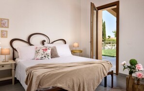 A bedroom with a bed, bedside lamps, a wooden headboard, a window with a view of greenery, and a door leading outside.