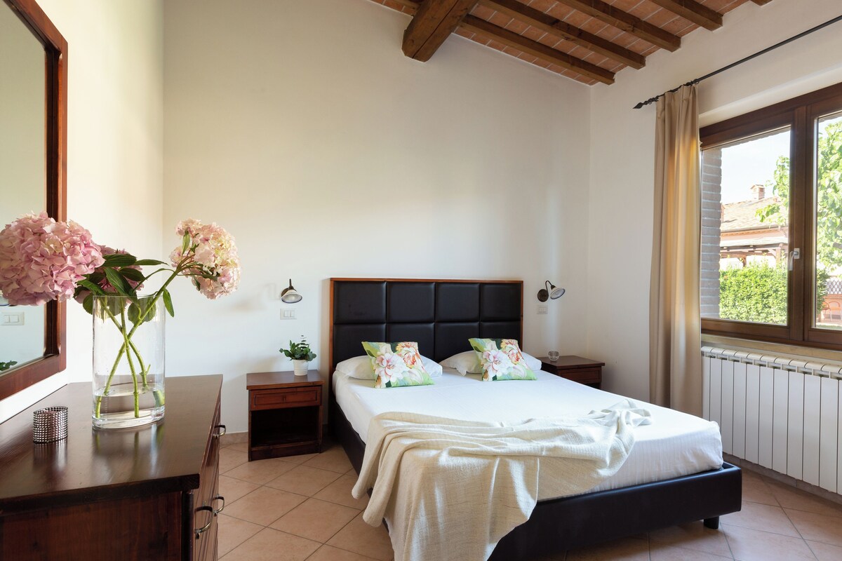 Une chambre à coucher avec un lit, des tables de chevet, un miroir, une fenêtre avec des rideaux et un vase de fleurs.