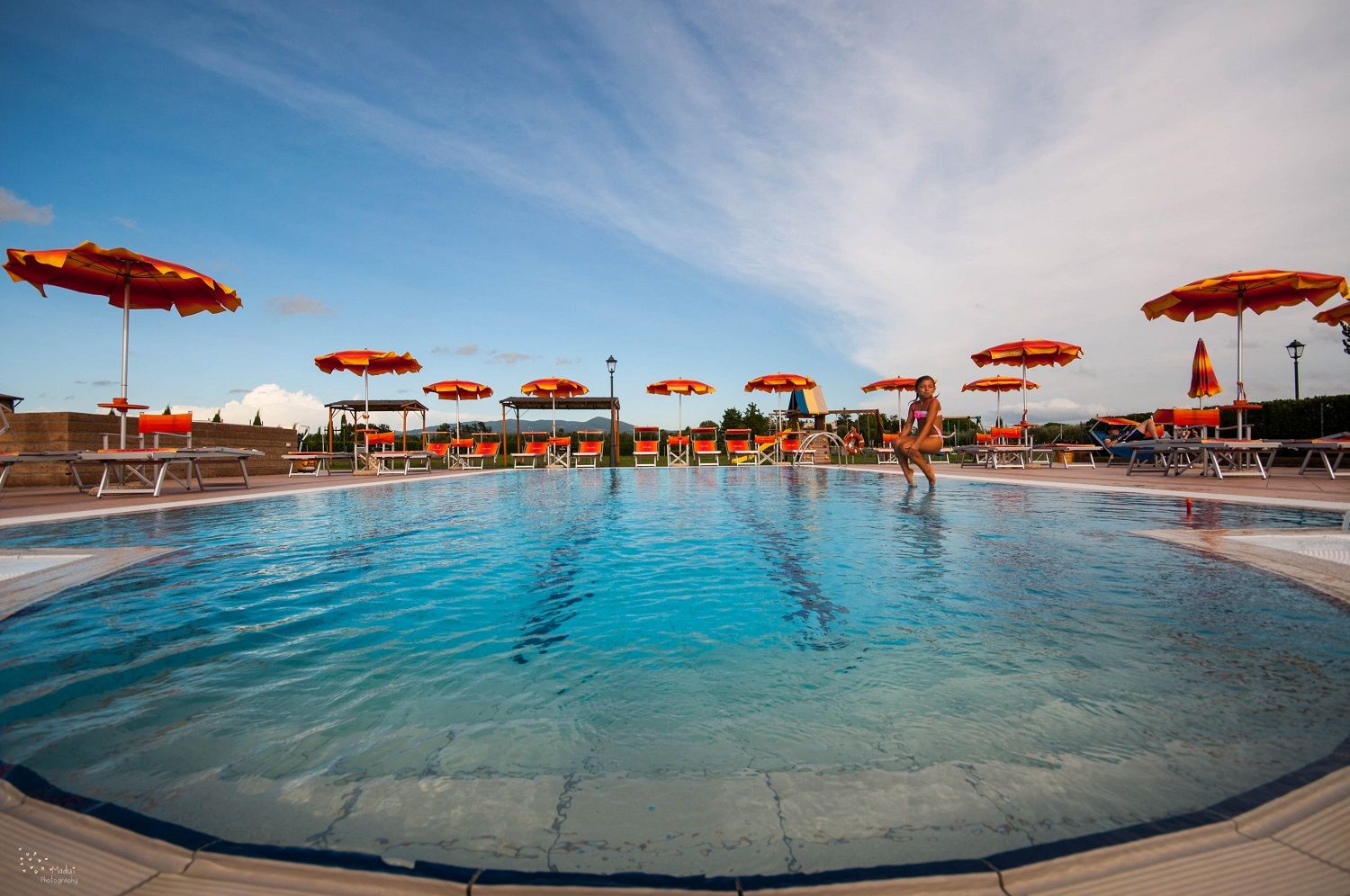 A swimming pool with multiple sun loungers and umbrellas, surrounded by a stone wall and a clear sky.