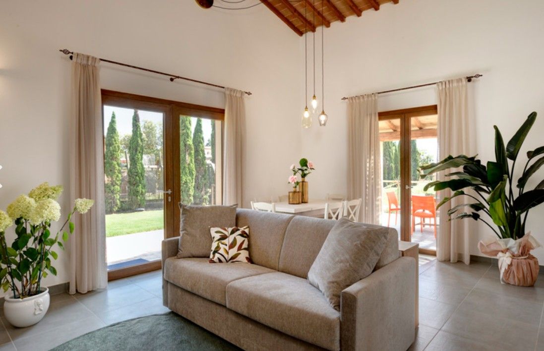 Un salon avec un canapé beige, une table basse et une grande plante en pot. Des rideaux sont accrochés aux fenêtres, et on peut voir le paysage à travers celles-ci.