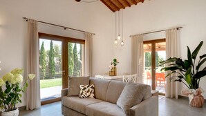 A living room with a beige sofa, a coffee table, and a large potted plant. There are curtains on the windows, and a view of the outdoors is visible. - Residence Borgo Verde (Rosignano Marittimo)