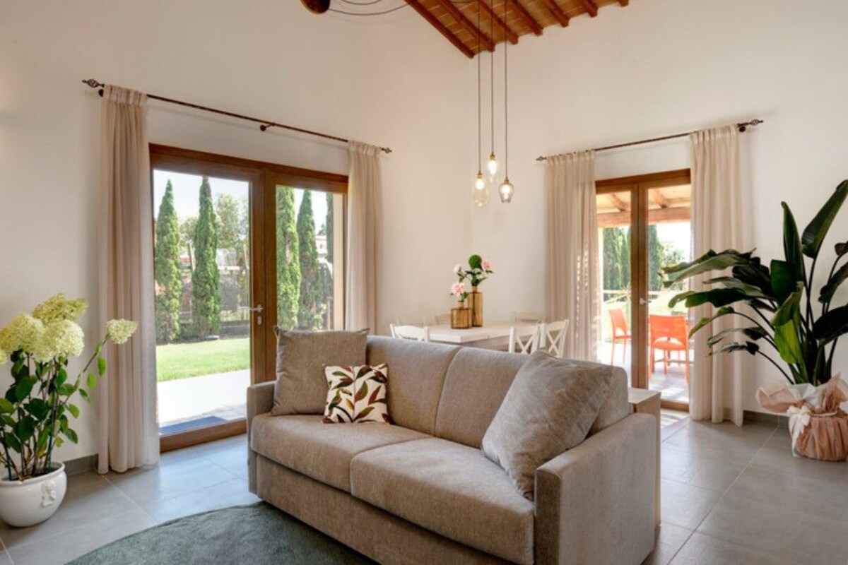 A living room with a beige sofa, a coffee table, and a large potted plant. There are curtains on the windows, and a view of the outdoors is visible.