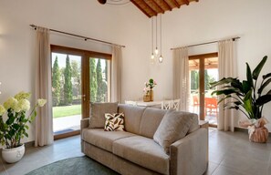 A living room with a beige sofa, a coffee table, and a large potted plant. There are curtains on the windows, and a view of the outdoors is visible. - Residence Borgo Verde (Rosignano Marittimo)