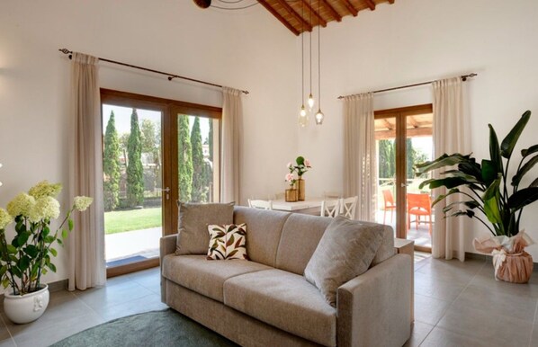 A living room with a beige sofa, a coffee table, and a large potted plant. There are curtains on the windows, and a view of the outdoors is visible. - Residence Borgo Verde (Rosignano Marittimo)