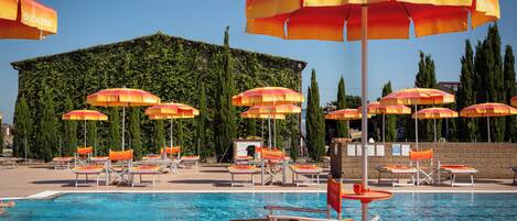 A pool area with multiple sun loungers and umbrellas, a clear blue pool, and a building with a green wall and trees in the background.