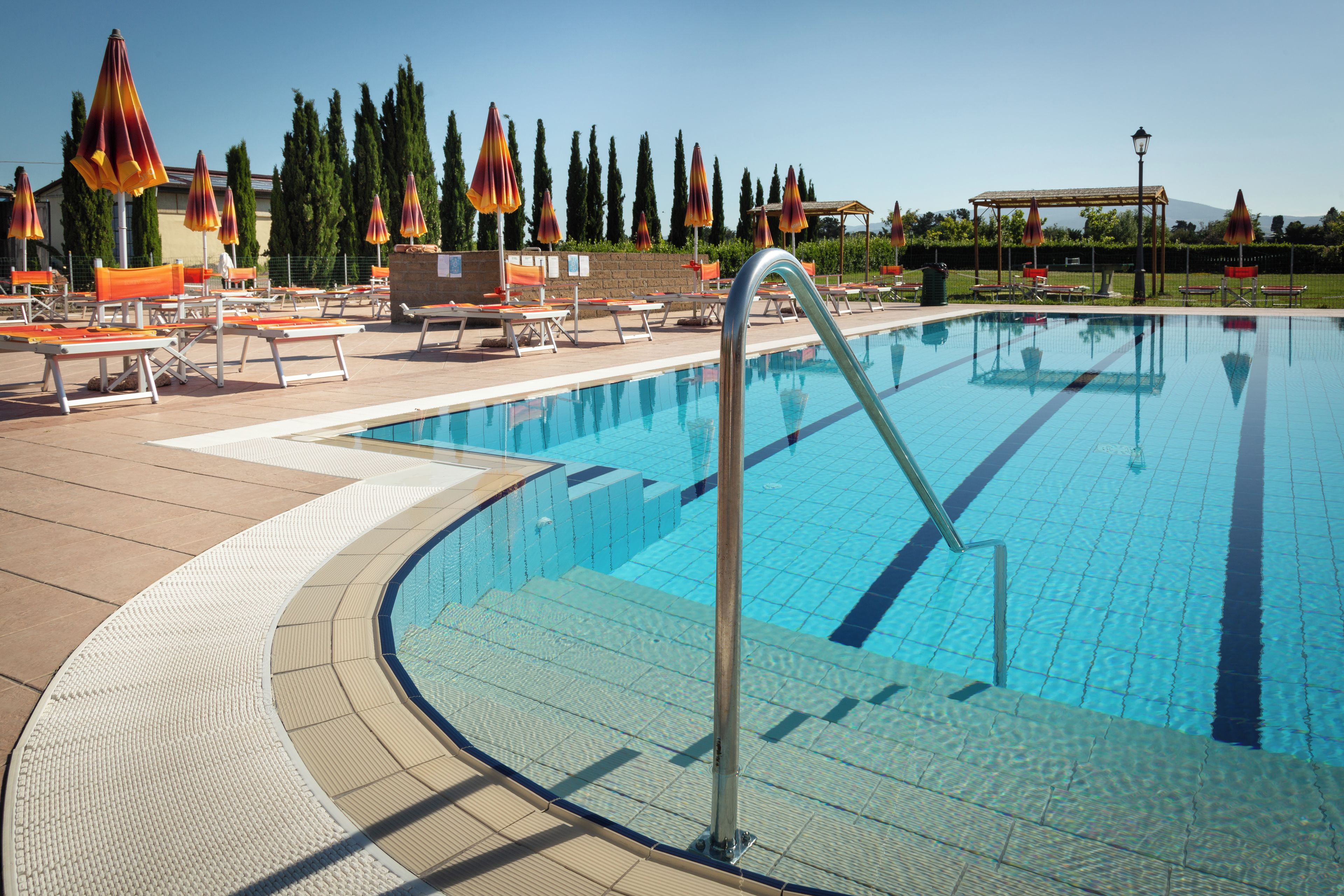 A swimming pool with sun loungers and umbrellas, surrounded by trees and a clear sky.