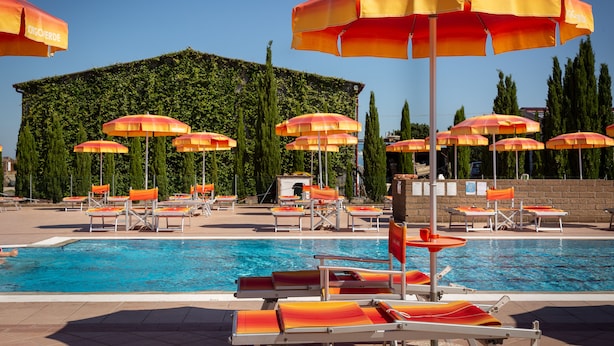 A pool area with multiple sun loungers and umbrellas, a clear blue pool, and a building with a green wall and trees in the background. - Residence Borgo Verde (Rosignano Marittimo)