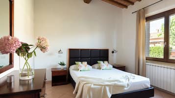 Un dormitorio con cama, mesitas de noche, un espejo, una ventana con cortinas y un jarrón con flores.