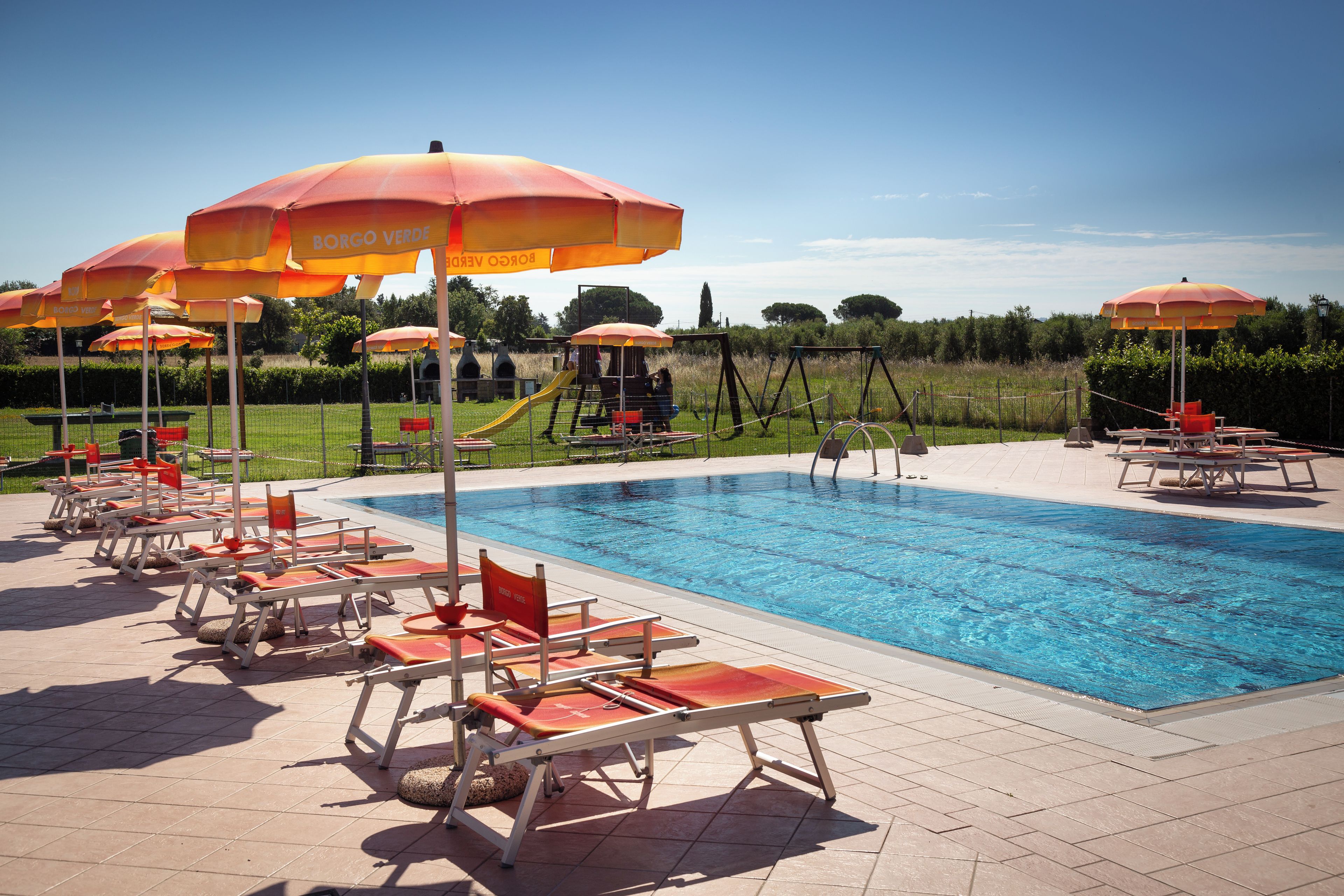 A swimming pool with sun loungers and umbrellas, surrounded by a fenced area with greenery.