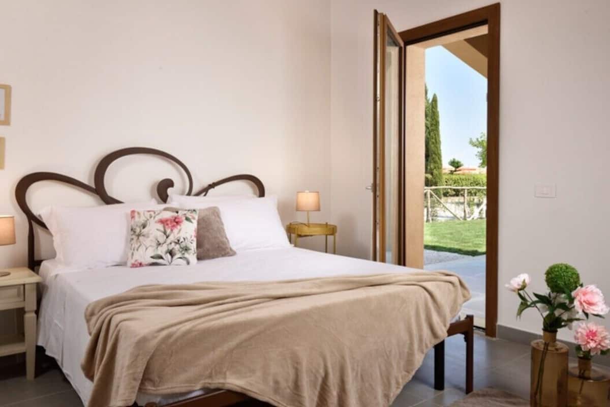 A bedroom with a bed, bedside lamps, a wooden headboard, a window with a view of greenery, and a door leading outside.