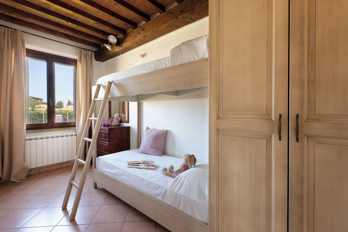 A bunk bed room with a ladder, a window with curtains, and a view of buildings outside.