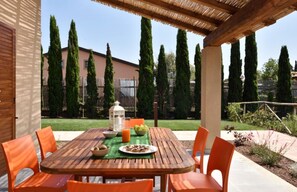 A wooden table with orange chairs under a pergola, set in a garden with tall cypress trees and a building in the background.