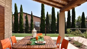 A wooden table with orange chairs under a pergola, set in a garden with tall cypress trees and a building in the background. - Residence Borgo Verde (Rosignano Marittimo)