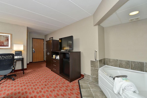 A hotel room with a red patterned carpet, a desk with a computer, a TV on a cabinet, and a white bathtub with towels. - Baymont by Wyndham Brooklyn Center (Brooklyn Center)