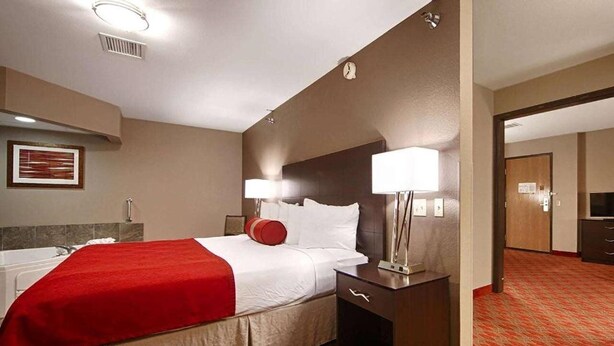 A hotel room with a bed, bedside table, lamp, and a clock on the wall. - Baymont by Wyndham Brooklyn Center (Brooklyn Center)