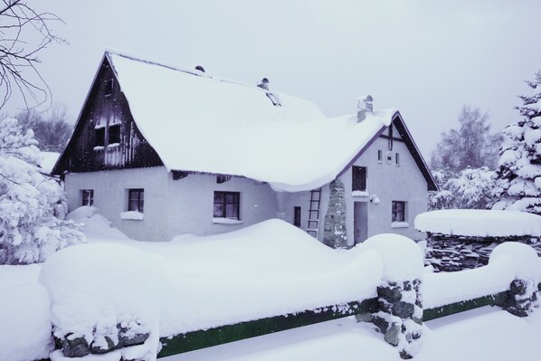 A snow-covered house with a steep roof, a visible chimney, and a stone wall in the foreground. - Chalupa na Fryšavě (Frysava pod Zakovou horou)