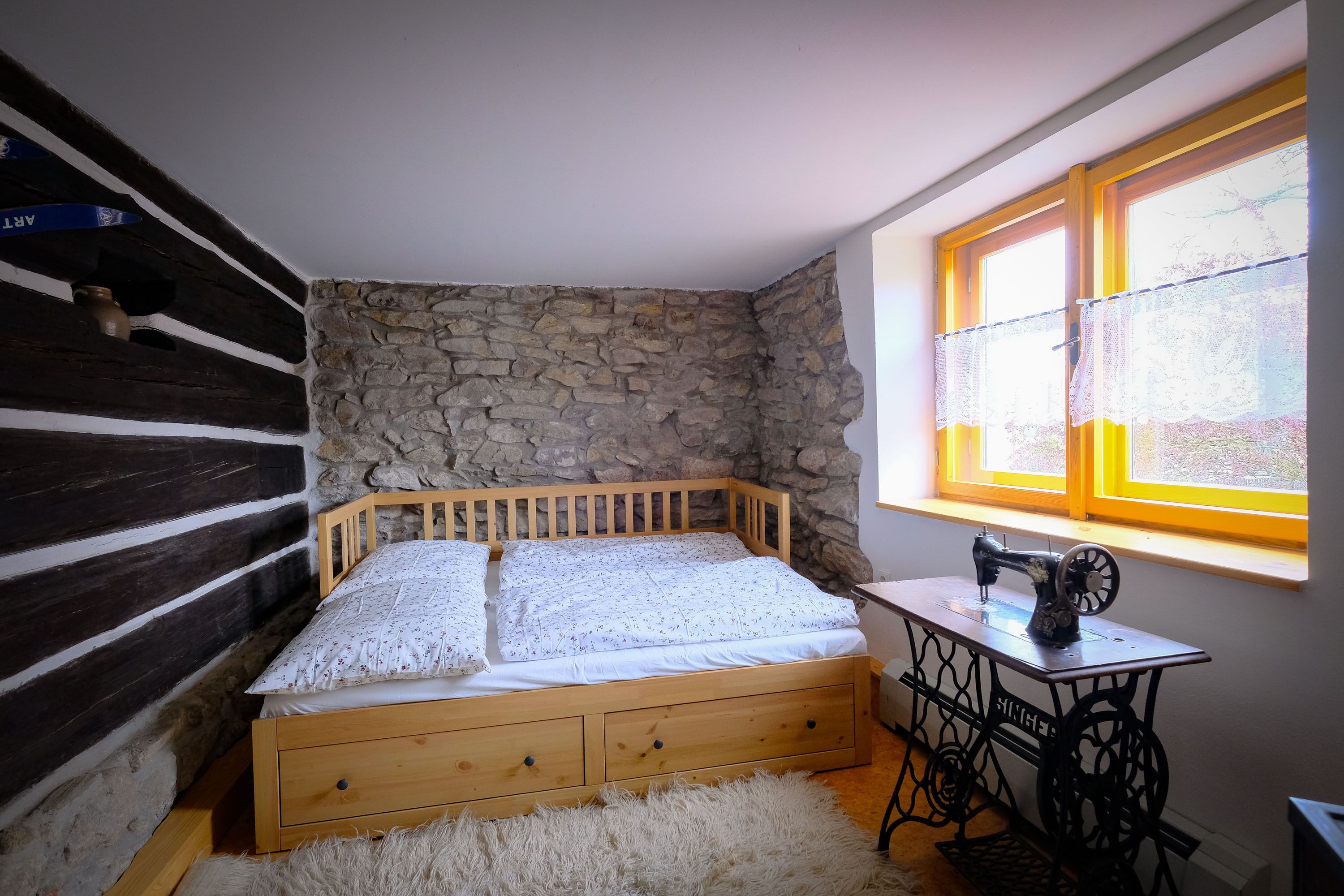 Une chambre avec un lit en bois, un mur en pierre et une machine à coudre près d’une fenêtre.