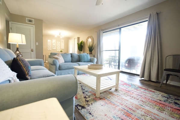 A living room with a sofa, armchair, and a coffee table. There is a window with curtains, a plant, and a lamp on a side table.
