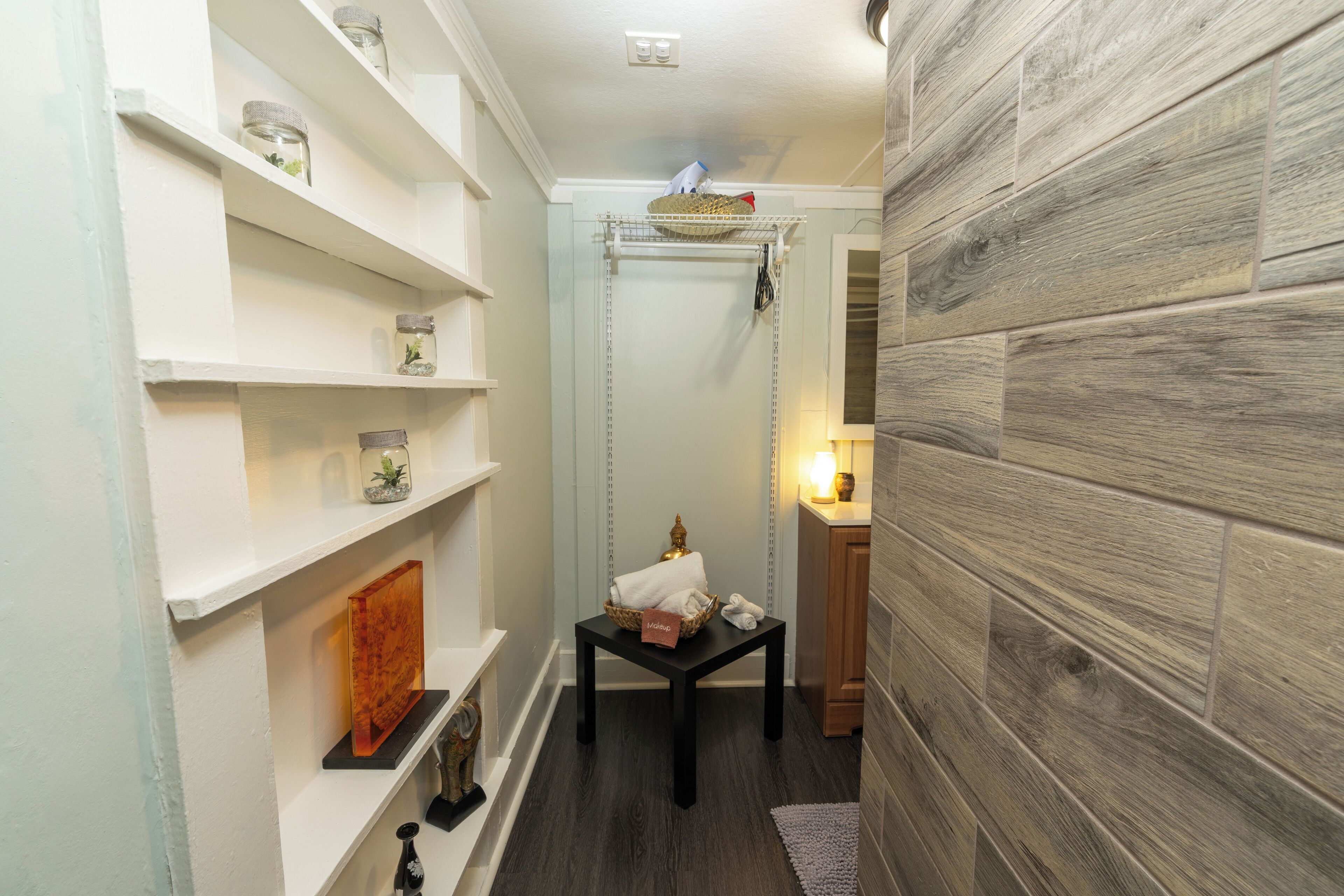 A narrow hallway with a wooden floor, white walls, and shelves with decorative items. There is a small table with a basket and a towel.