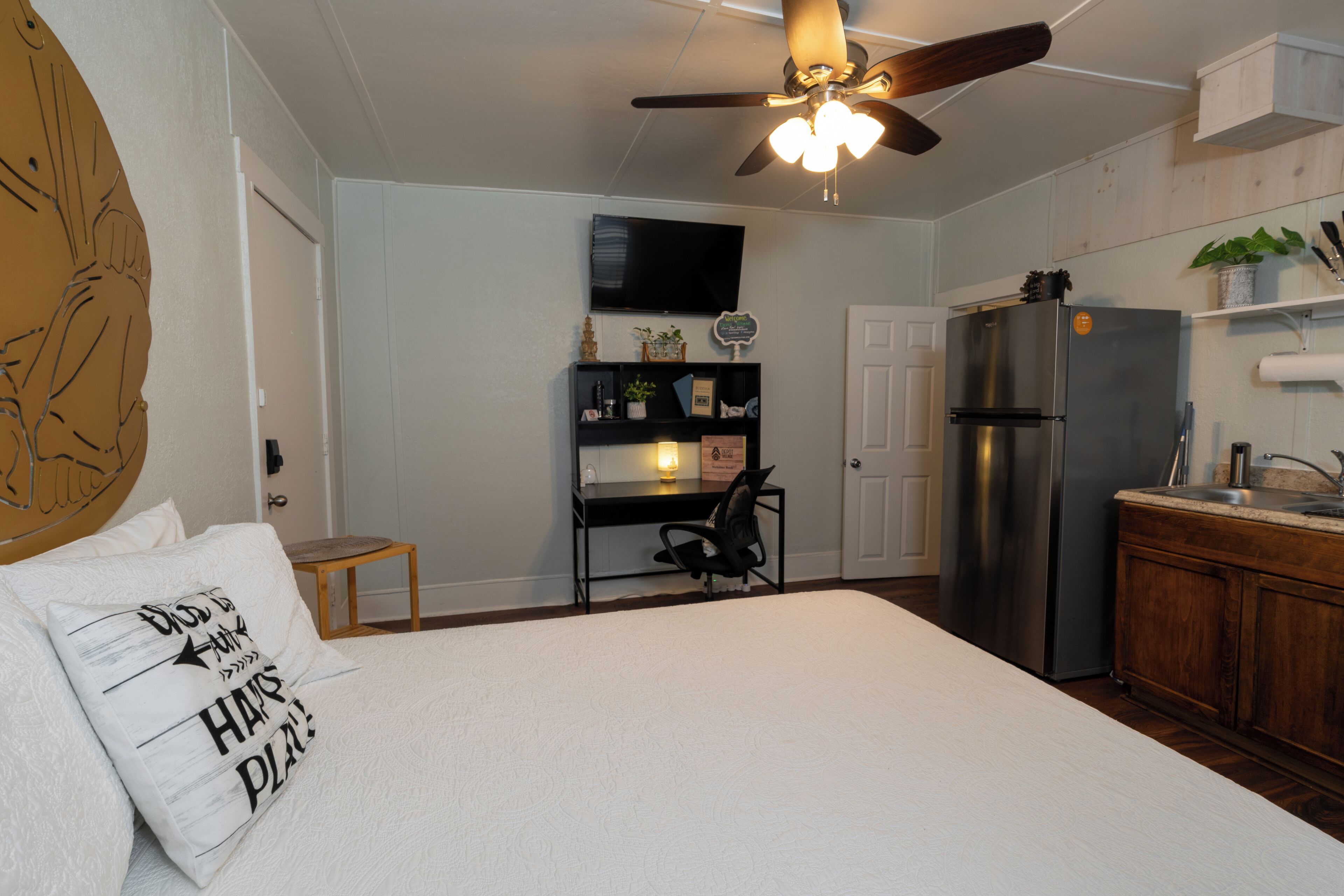 Une chambre comprenant un lit, un bureau, une chaise, un ventilateur de plafond, un réfrigérateur et un coin cuisine avec un évier et des placards.