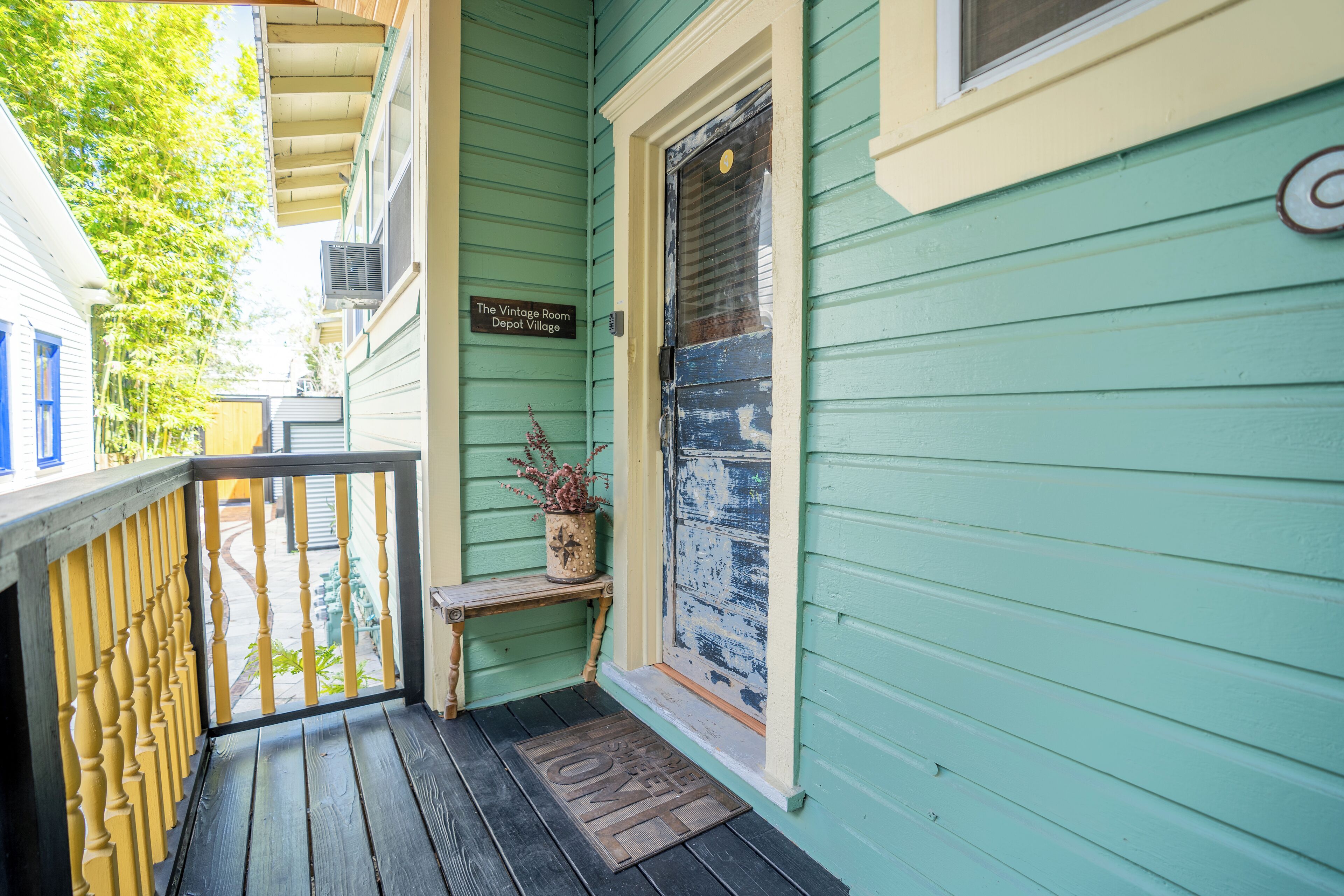 A porch with a wooden bench, a potted plant, and a door with a sign reading "The Vintage Room Depot Village".