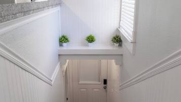 A white interior hallway with a marble staircase, a door with a window, and three potted plants on a shelf.