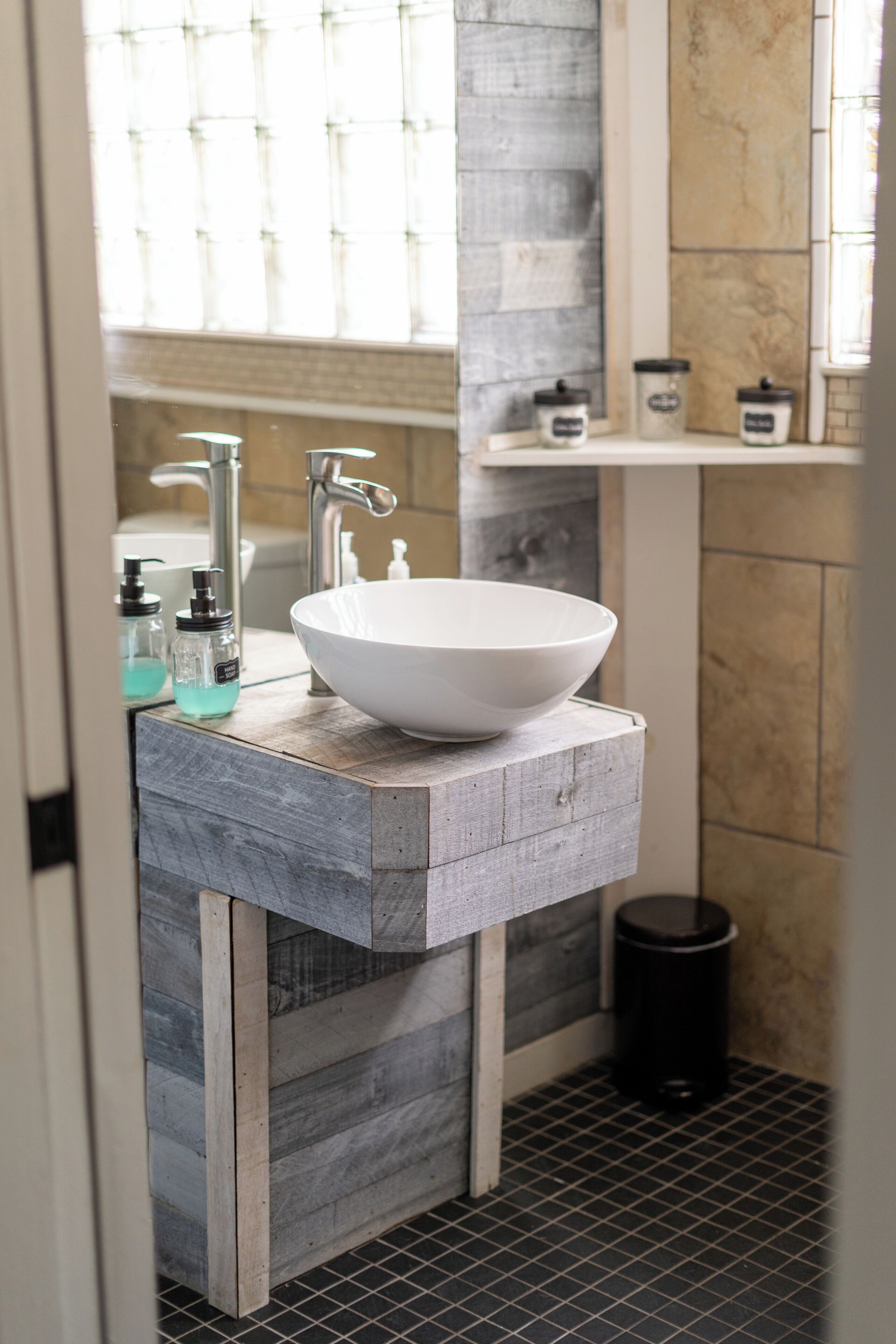 A bathroom with a large white sink, a wooden vanity, a black trash can, and a window with blinds.