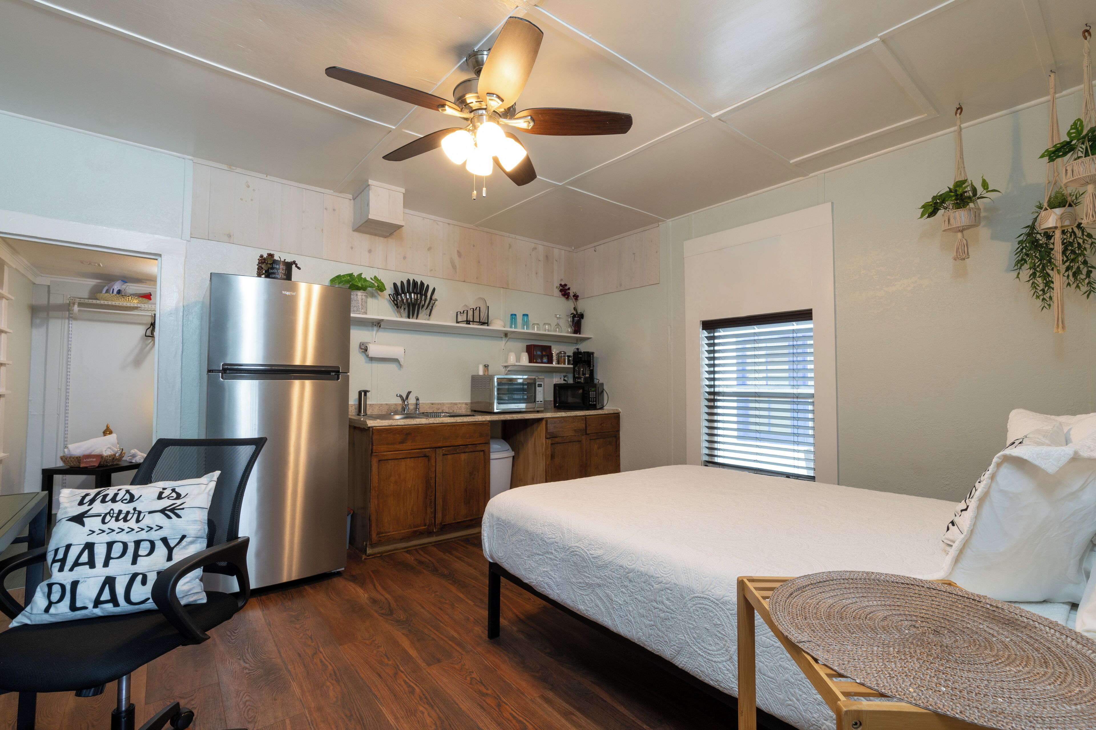 A compact kitchen with a refrigerator, microwave, and sink. There is a bed with a white comforter and a ceiling fan.