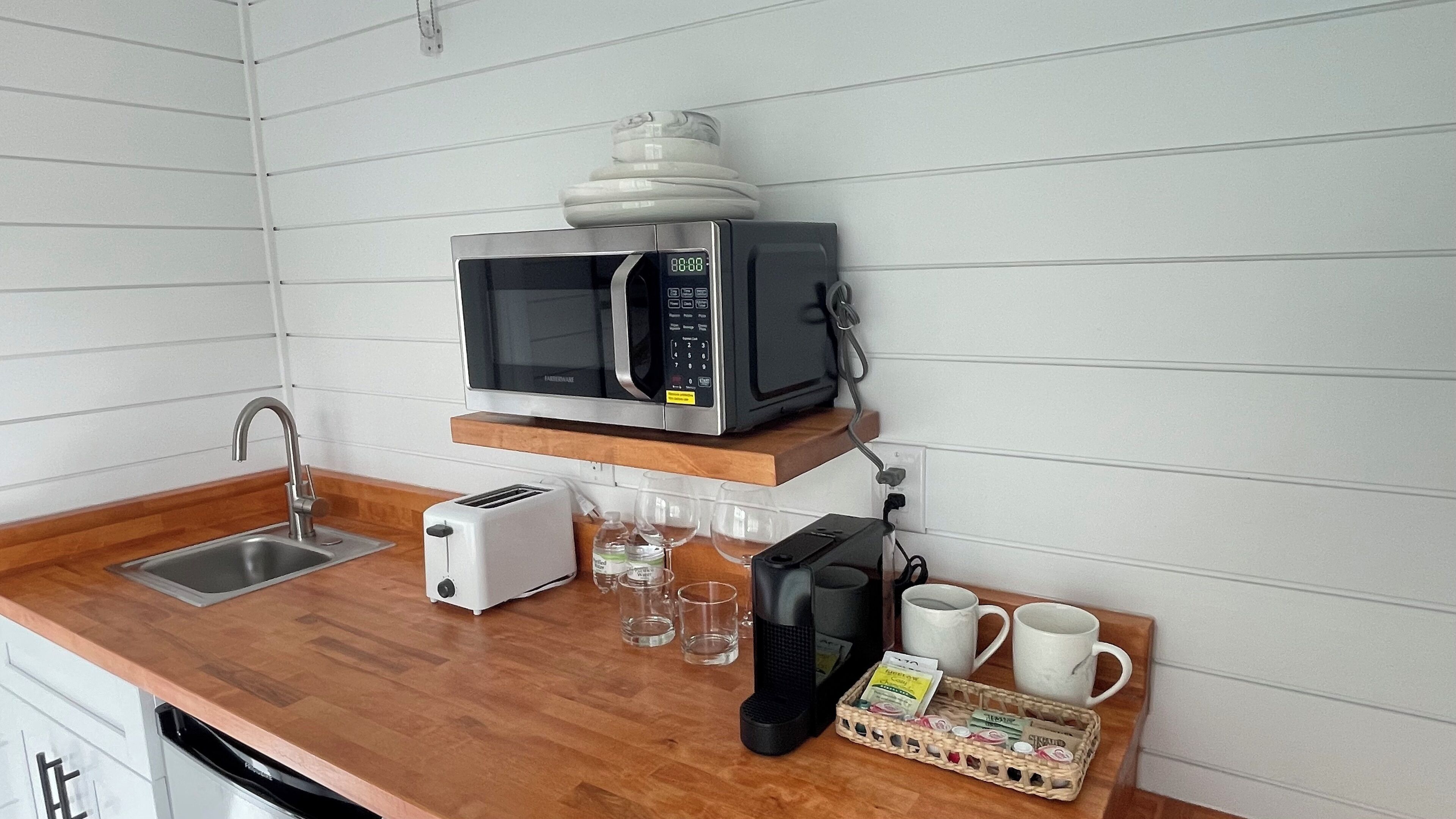 A kitchen with a microwave, toaster, sink, and a coffee maker. There are also wine glasses and a basket of tea bags.