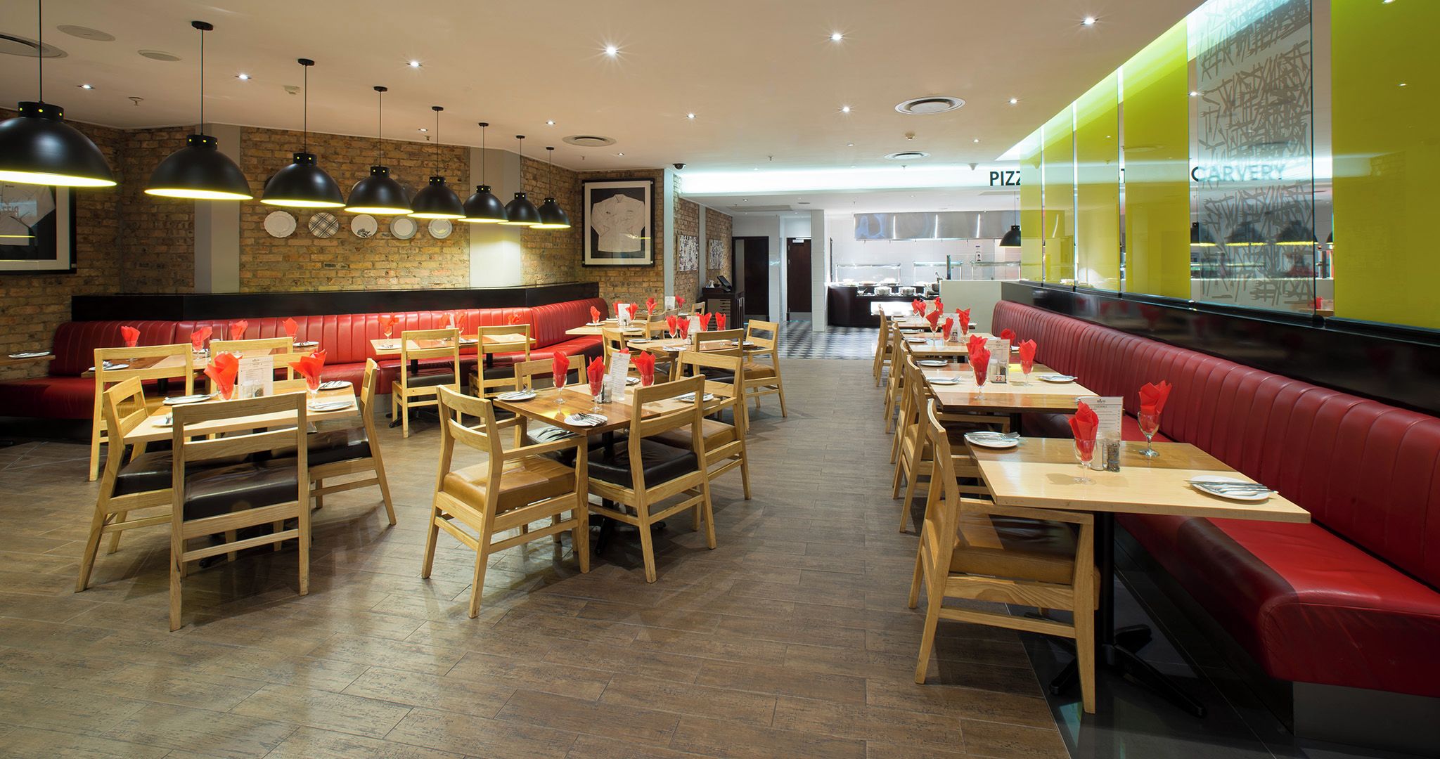 A restaurant with wooden tables and red upholstered booths, featuring a brick wall and pendant lighting.
