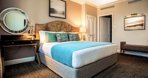A hotel room with a large bed, a round mirror, a bedside table, a lamp, a bench, and a door. - The Ridge Hotel (Emalahleni)