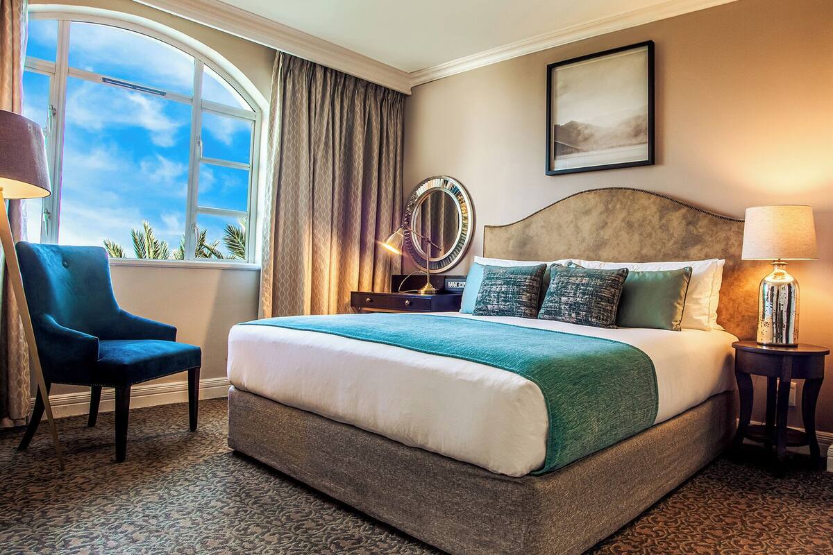 A hotel room with a large bed, a chair, a bedside table with a lamp, a mirror, and a door.