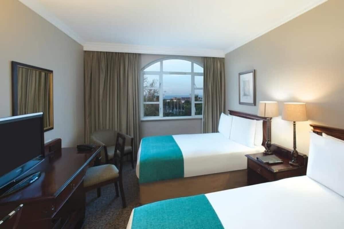 A hotel room with two beds, a desk, a television, and a large window with curtains.