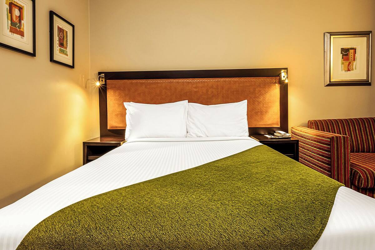 A hotel room with a large bed, a green throw blanket, a striped armchair, and framed artwork on the walls.