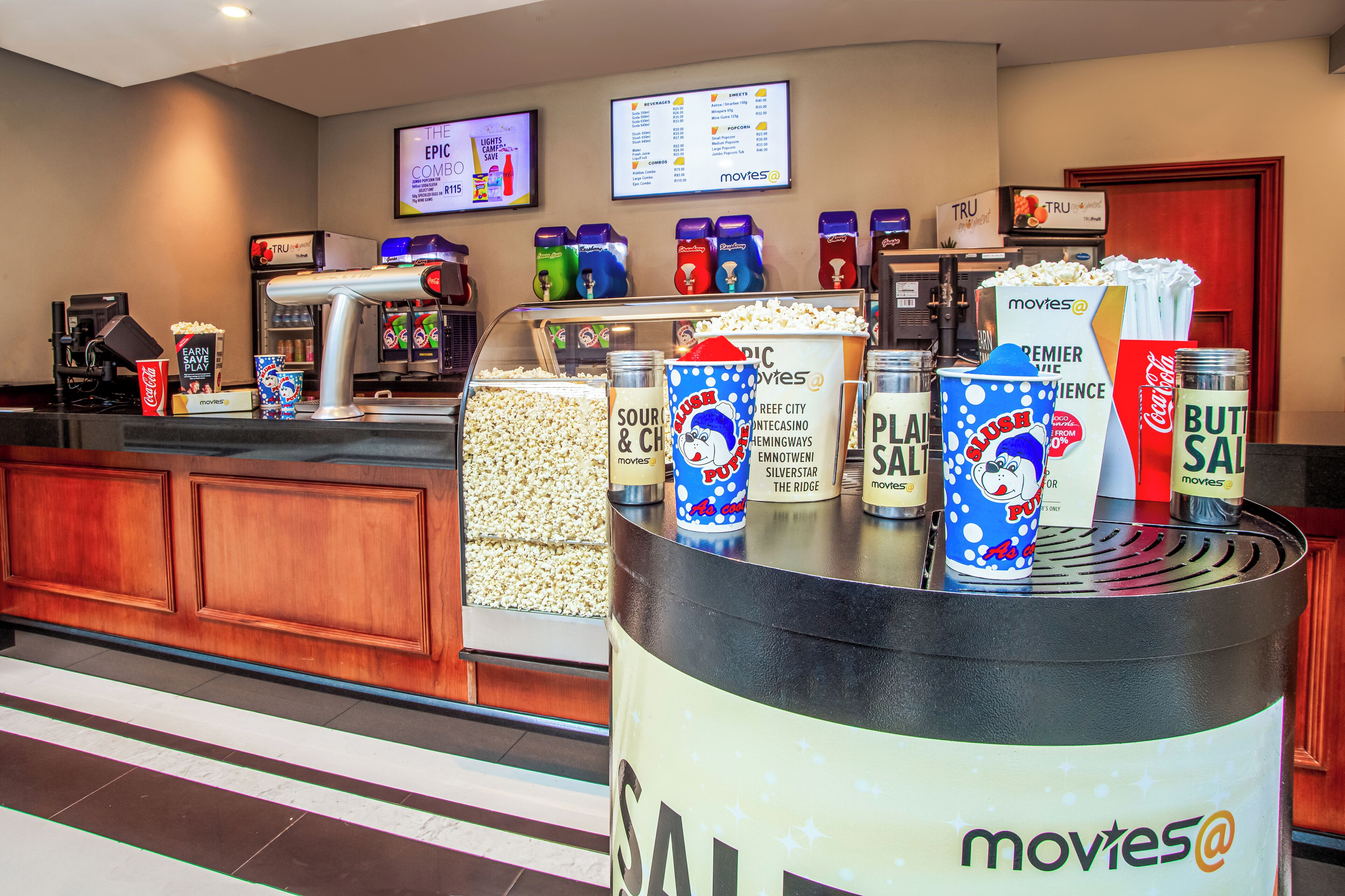 A cinema lobby with popcorn and drink dispensers, a popcorn box, and a menu board.