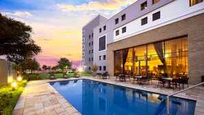 A swimming pool with a patio area, outdoor seating, and a building with large windows. - The RidgePoint Hotel (Emalahleni)