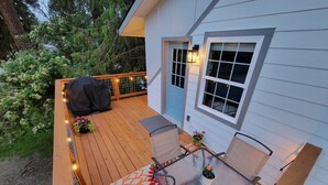 Una terraza de madera con mobiliario exterior, una parrilla cubierta y luces colgantes.