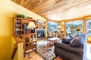 Un salon chaleureux avec une cheminée, des étagères à livres, un canapé et de grandes fenêtres offrant une vue sur la montagne.