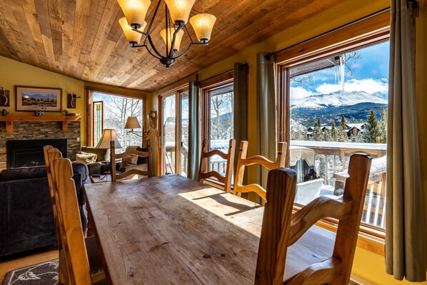 Une pièce spacieuse avec une grande table en bois, des sièges confortables, une cheminée et de grandes fenêtres donnant sur une vue imprenable sur les montagnes.