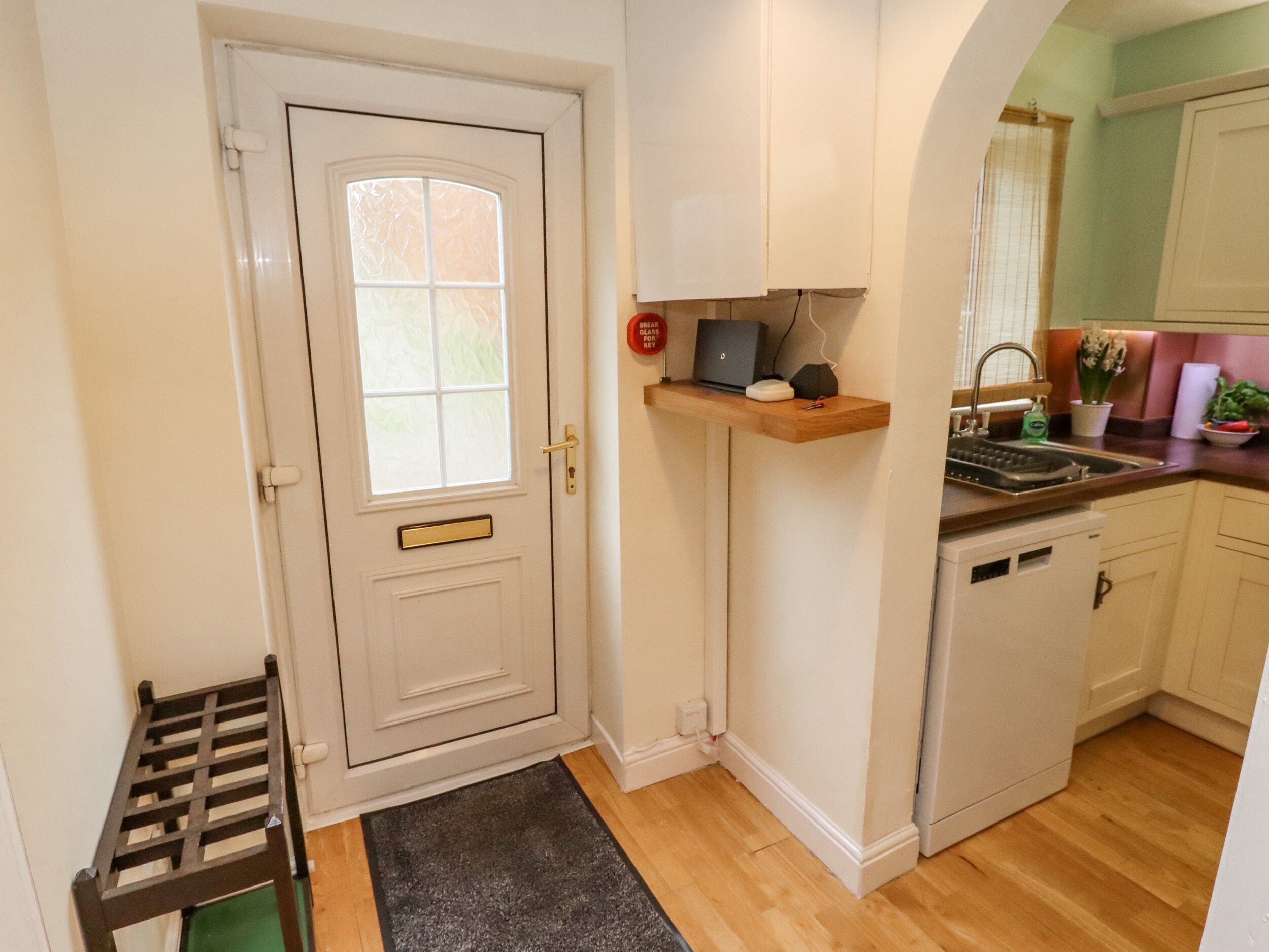A compact kitchen with a white door, a small window, a wooden shelf with a laptop, a dishwasher, and a green mat at the entrance.