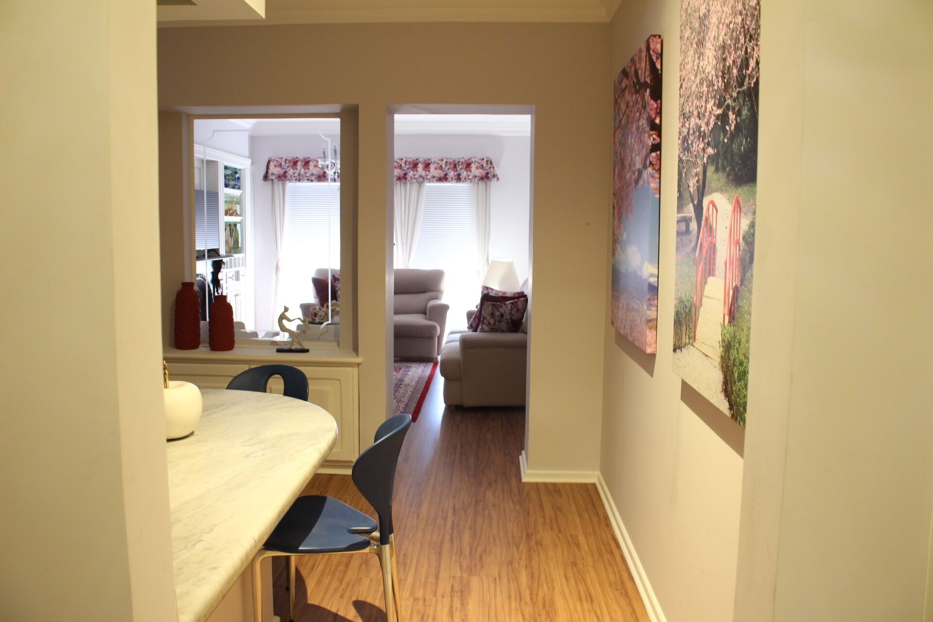 A hallway with a dining area on the left, a living room with a sofa and a lamp on the right, and a painting on the wall.