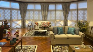 A spacious living room with a beige sofa, a coffee table, and a decorative rug. There are two windows with curtains, a small Christmas tree, and a lamp on a side table.