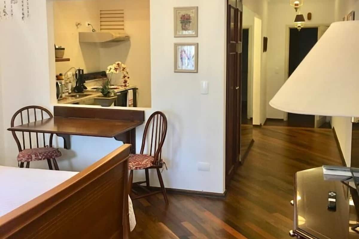 A spacious living area with a wooden dining table, two chairs, a kitchen in the background, and a hallway leading to another room.