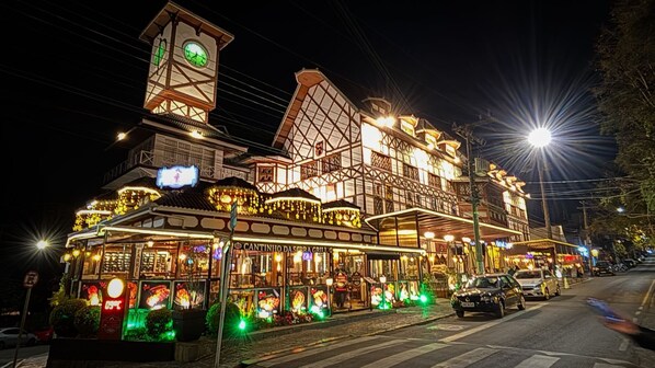 A brightly lit building with a prominent clock tower, multiple windows, and a sign reading "Cantinho da Serra Grill". - Center Suíço Flats e Shopping (Campos do Jordão)