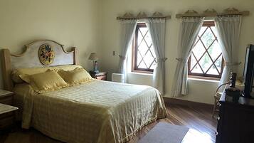 A bedroom with a bed, two windows with curtains, a wooden headboard, a bedside table, a lamp, and a dresser.