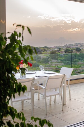 A dining table with chairs and a vase of flowers, set against a scenic view of hills and a sunset. - A52.mt - Apartment with Sea & Country Views (Mellieha)